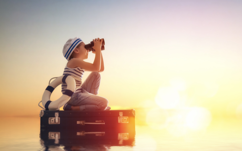 A child sits atop a suitcase, donning a sailor's hat and binoculars, gazing out at the vast ocean as the sun sets on the horizon. This whimsical image embodies the idea of 'coming into union with your true self' and the joy and wonder that comes with manifesting your dreams and setting sail on the journey of a lifetime.